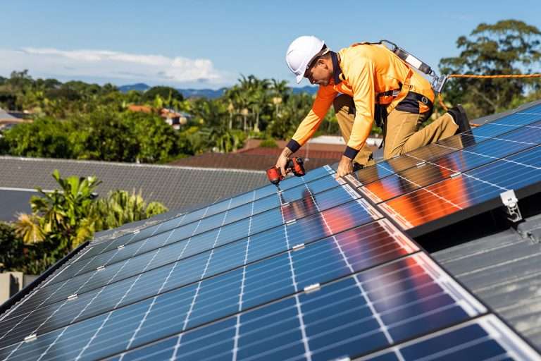 Solar panel technician with drill installing solar panels on house roof on a sunny day.