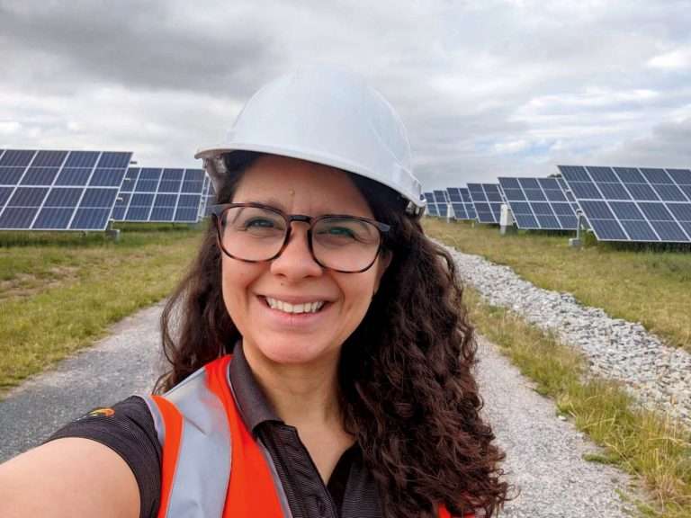 Woman in a field of solar panels, wearing a white hard hat and hi-vis vest