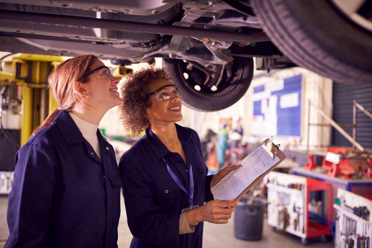 Two women looking at the underside of a car.