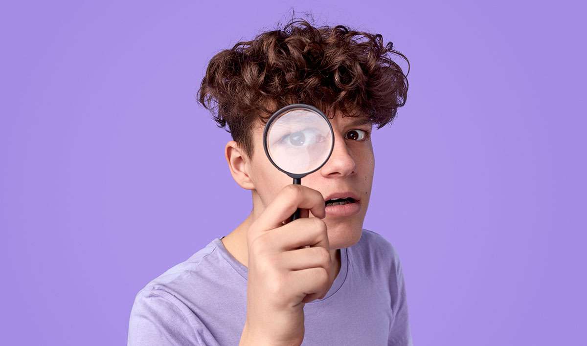 Curious teen boy with looking at camera through magnifying glass while searching for clues against violet background
