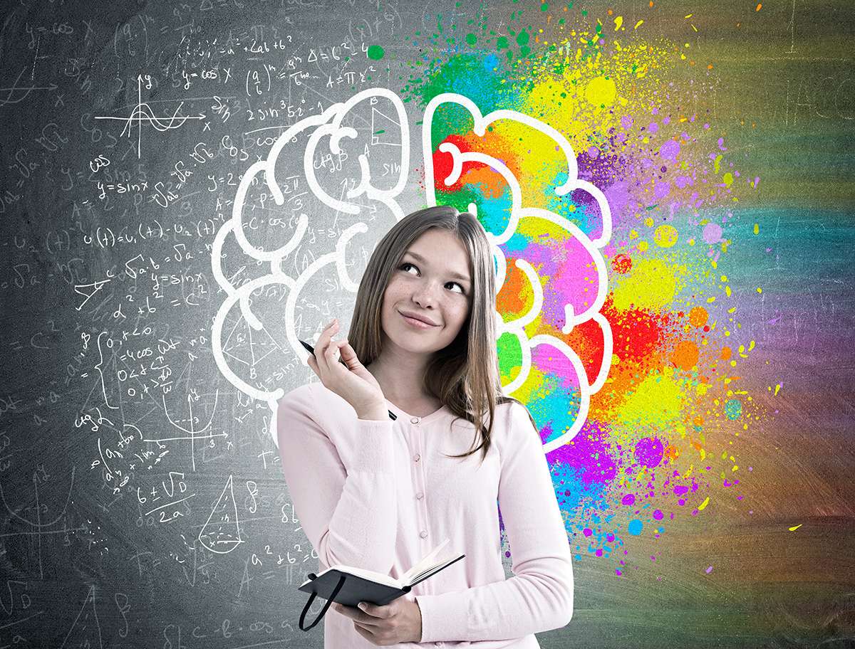 Portrait of a dreamy girl in a pink sweater holding a planner and a pen and standing near a blackboard with a colorful brain sketch and formulas on it.