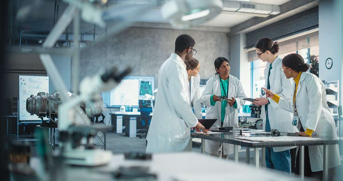 Wide Shot of Group of Young Multiethnic Lab Specialists Working as a Team to Analyse Blueprints and Digital Components Using Laptop Computer and Tablet. Professional Scientists Brainstorming