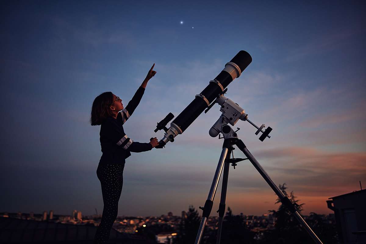 Girl with astronomical telescope stargazing under twilight sky.