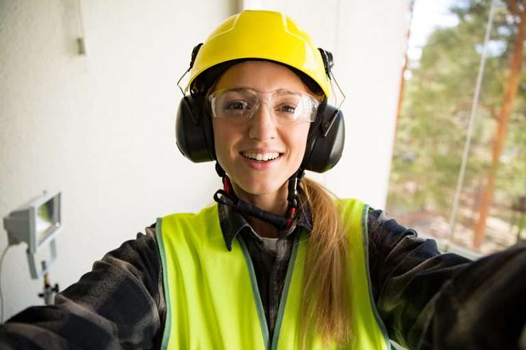 Portrait of a Young Female construction worker in hard hat drilling concrete wall with a drill and smiling at the camera. Building and renovation. Selfie.