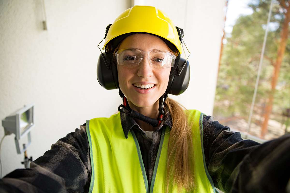 Portrait of a Young Female construction worker in hard hat drilling concrete wall with a drill and smiling at the camera. Building and renovation. Selfie.