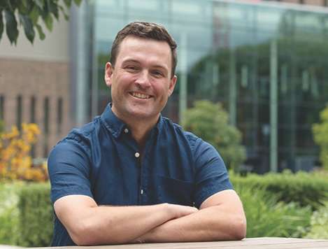 Man in blue shirt with a building and bushes in the background