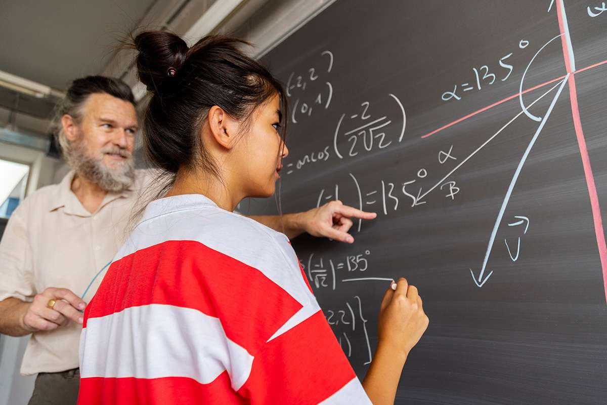 High school maths teacher explains blackboard exercise to teen student.
