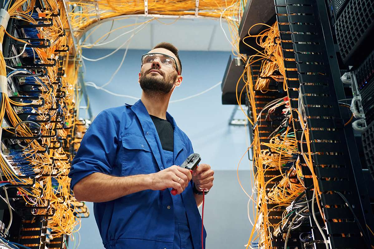 Young man is working with internet equipment and wires in server room.