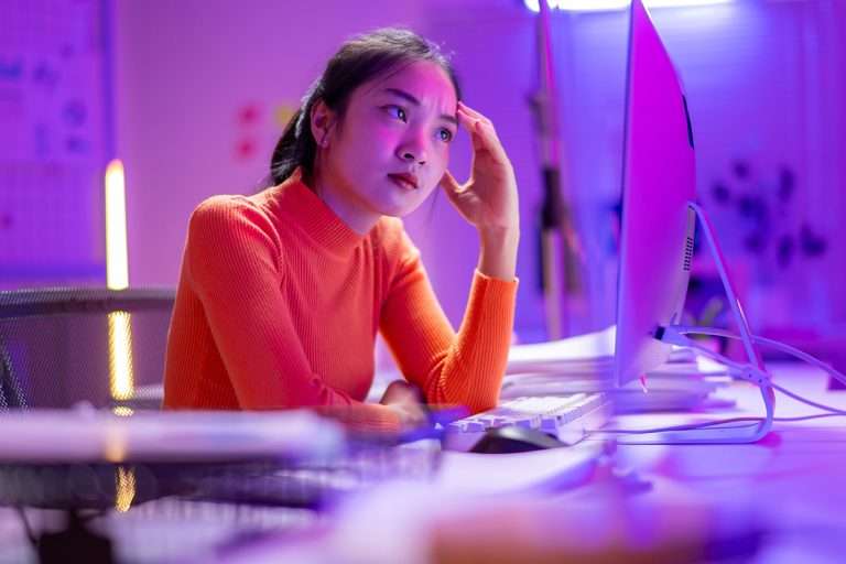A woman in an orange shirt is sitting at a computer desk with a keyboard and mouse in front of her. She is looking at the computer screen with a frown on her face
