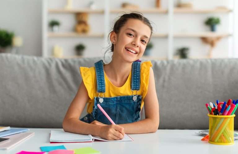 Girl sitting at table, writing and posing at camera