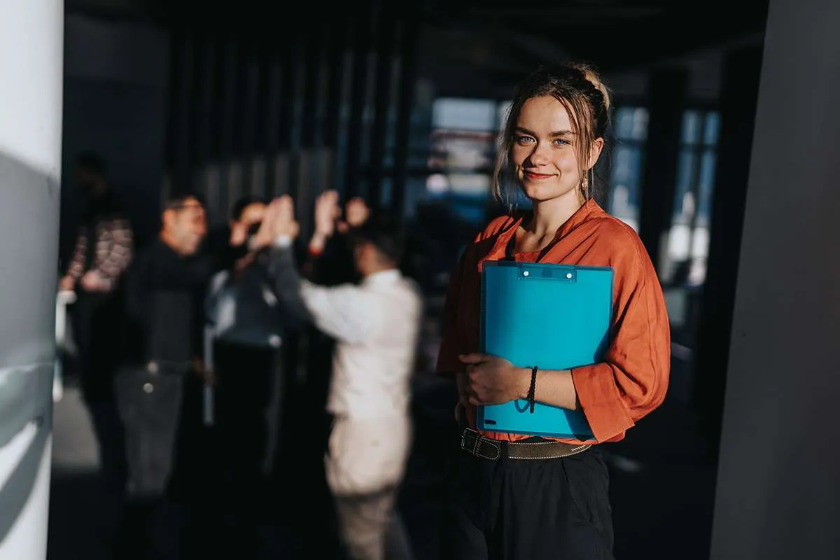 A confident female social scientist stands holding a blue clipboard, with colleagues interacting energetically in the background. The image captures teamwork and leadership in a modern office setting.